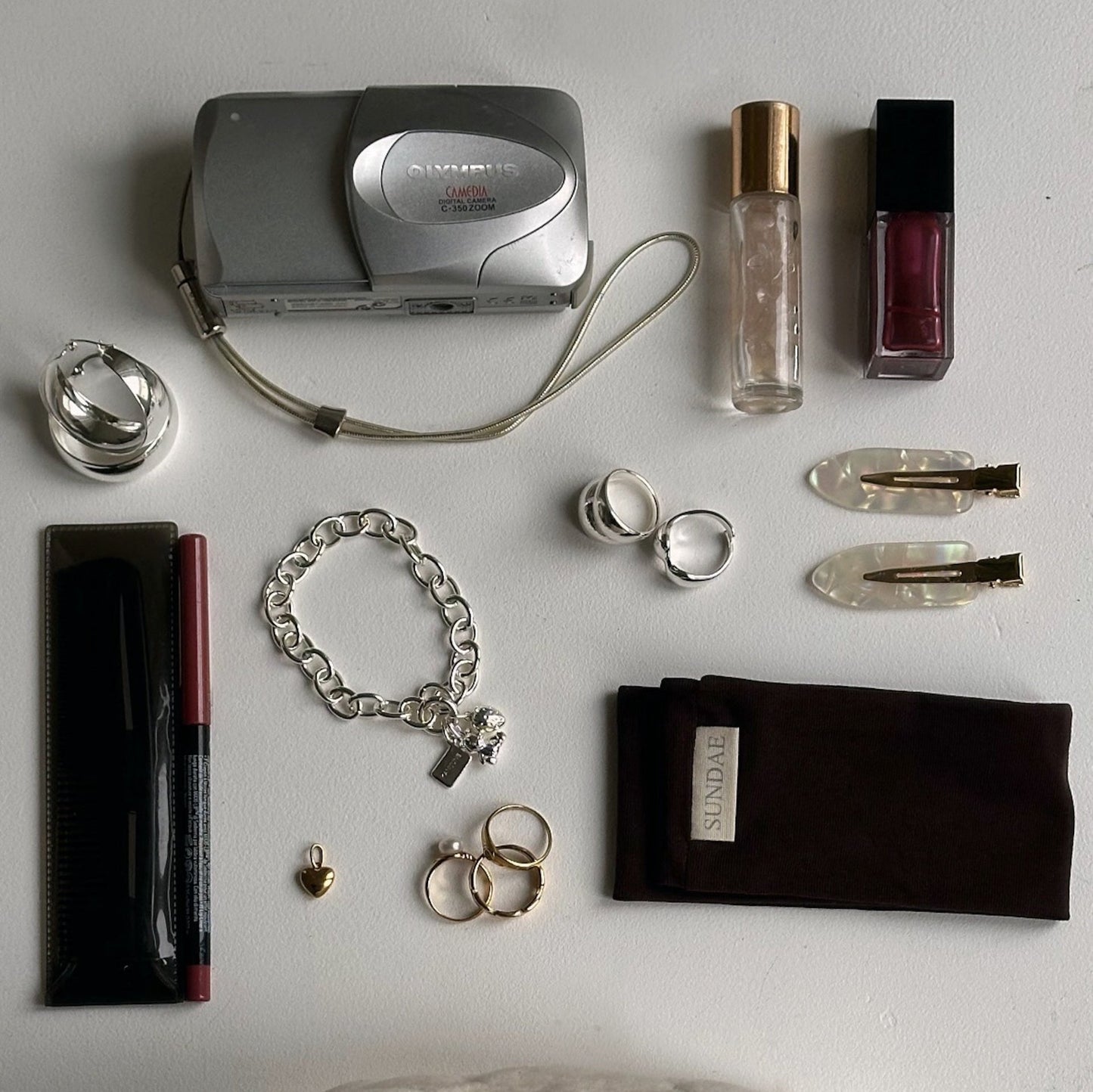 constellation of camera, lip gloss, make up and hair products staged on white desk with jewelry pieces in silver