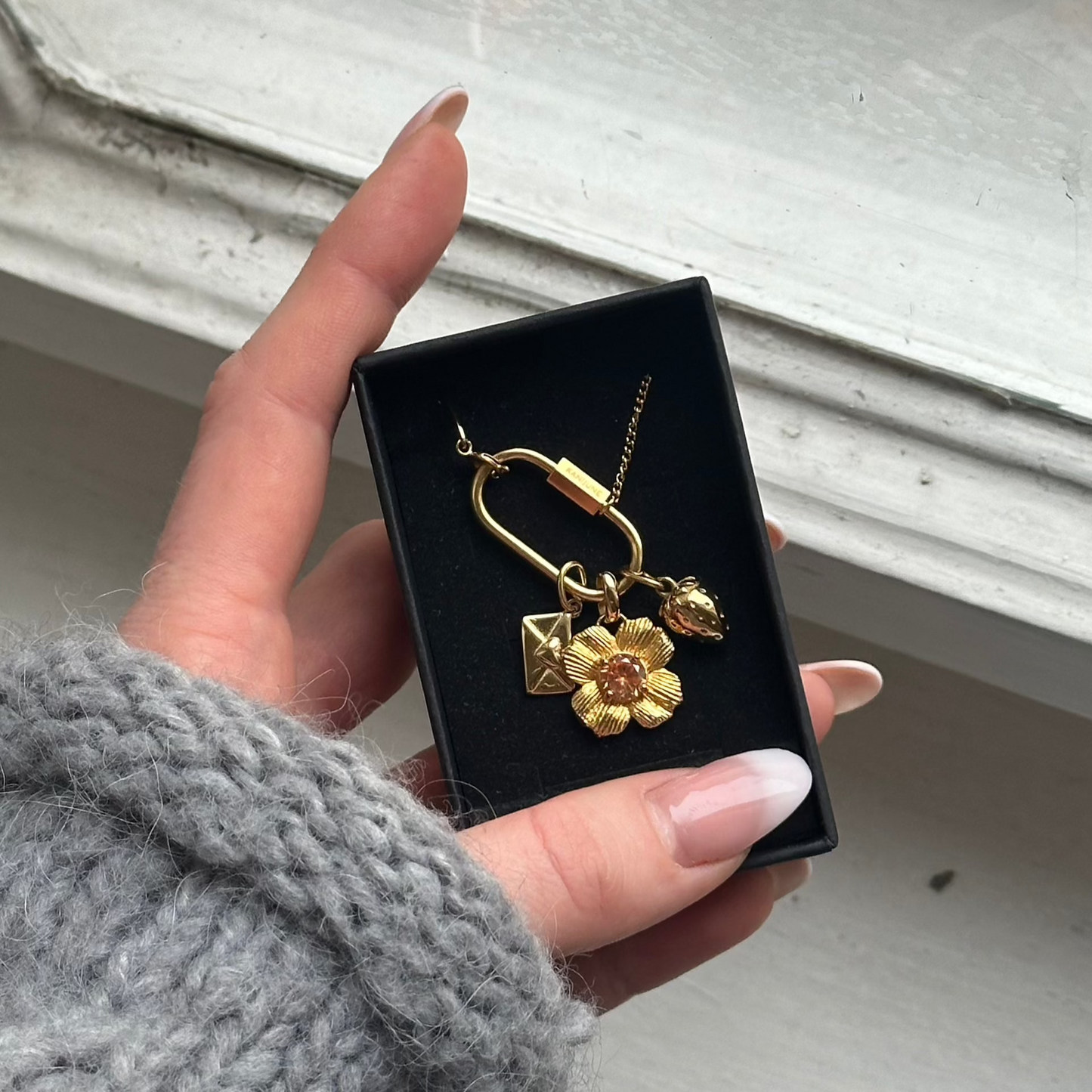 mix and match necklace with love letter, champagne flower and strawberry charm in jewelry box hold by a hand in fron of which wood and a glass window